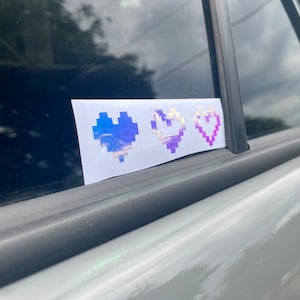 Puede incluir: Una pegatina con tres corazones pixelados en tonos azules, blancos y morados. La pegatina está adherida a la ventana de un coche. Los corazones están dispuestos horizontalmente y tienen un acabado holográfico iridiscente.