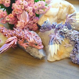 Pu&ograve; includere: Due piccole statuine di drago decorative fatte di fiori di stoffa rosa e viola. I dragoni sono posizionati su rocce bianche con un'arrangiamento di fiori rosa sullo sfondo.