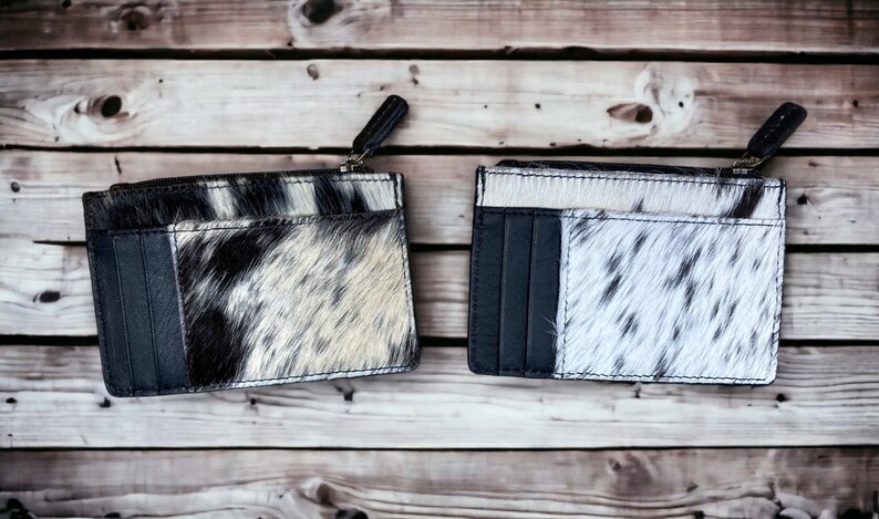 May include: Two black and white cowhide leather card holders with a zipper closure. The card holders have multiple card slots and are made of a soft, supple leather.