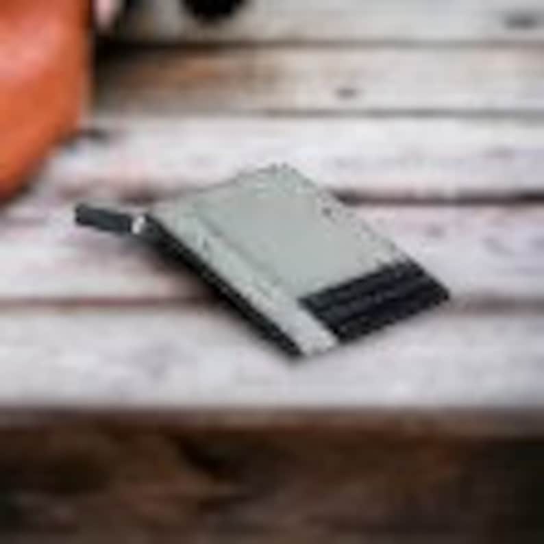 May include: A gray minimalist wallet with a black elastic band to hold cards. The wallet has a metal clip on the side.