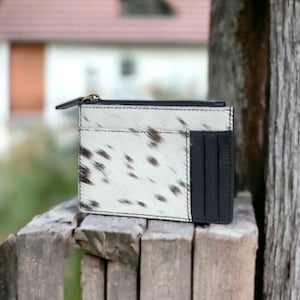 May include: A white and black cowhide leather wallet with a zipper closure and a black leather card slot.