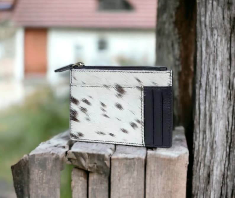 May include: A white and black cowhide leather wallet with a zipper closure and a black leather card slot.
