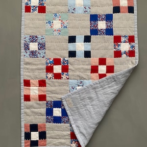 Puede incluir: Una colcha de retazos con un fondo gris claro y cuadrados de tela roja, azul y blanca. La colcha está doblada por la mitad y el reverso es de tela azul claro.