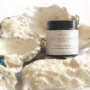 Puede incluir: Un tarro blanco con tapa negra y etiqueta que dice "VIA MERULANA ORGANICS PEARL CREAM Nano-meter Pearl Powder - Tallow - Castor Oil". El tarro está sobre una pila de conchas blancas.