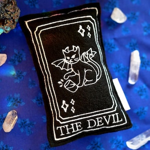 May include: Black rectangular tarot card with white embroidered outline and text "THE DEVIL". The card features a devil cat illustration. The card is surrounded by crystals and sits on a blue floral fabric.