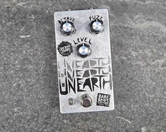 以下が含まれることがあります： テクスチャ仕上げのシルバーのギターエフェクトペダル。Octave、Fuzz、Levelとラベル付けされた3つの黒いコントロールノブを備えています。ペダルには、Sketchy Sounds、Unearth、Bare Bones Editionという文字がスタイリッシュな黒いフォントで記されています。
