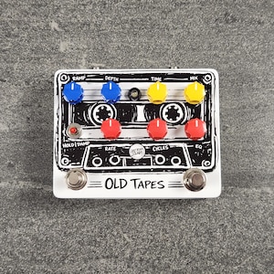 May include: White guitar effects pedal with a black and white cassette tape design. It has blue, yellow, and red knobs, and the text "OLD TAPES" is printed on the bottom. The pedal has two silver foot switches.