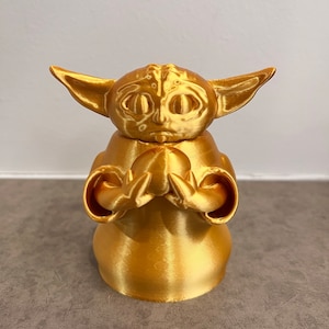 K&ouml;nnte beinhalten: Eine goldfarbene 3D-gedruckte Figur eines Charakters aus dem Star Wars-Franchise, bekannt als "The Child" oder "Baby Yoda". Die Figur h&auml;lt ein kleines, rundes Objekt in ihren H&auml;nden.
