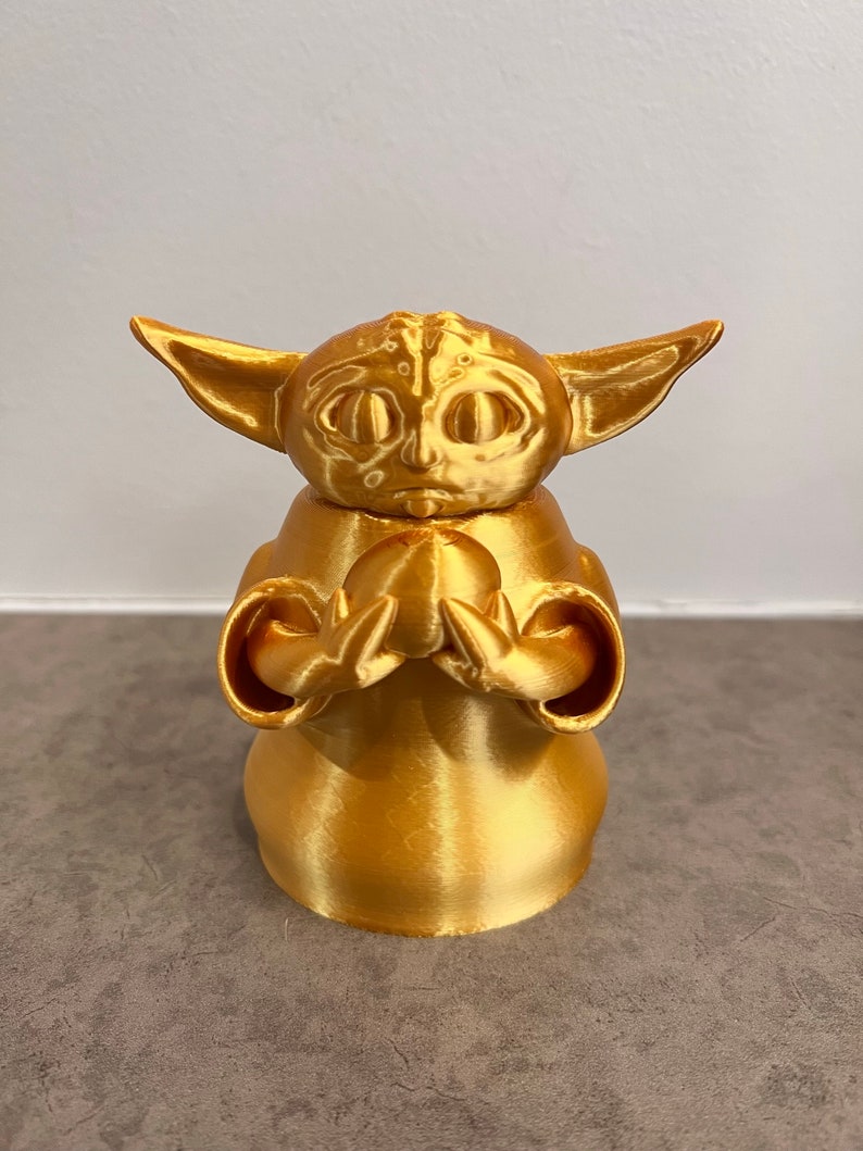 K&ouml;nnte beinhalten: Eine goldfarbene 3D-gedruckte Figur eines Charakters aus dem Star Wars-Franchise, bekannt als "The Child" oder "Baby Yoda". Die Figur h&auml;lt ein kleines, rundes Objekt in ihren H&auml;nden.