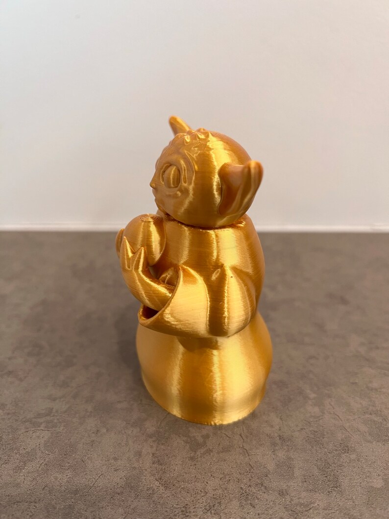 K&ouml;nnte beinhalten: Eine goldfarbene 3D-gedruckte Figur eines beliebten fiktiven Charakters. Die Figur h&auml;lt ein kleines Objekt in ihren H&auml;nden.