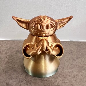 K&ouml;nnte beinhalten: Eine goldfarbene 3D-gedruckte Figur eines Charakters aus dem Star Wars-Franchise, bekannt als "The Child" oder "Baby Yoda". Die Figur ist mit ineinander verschr&auml;nkten H&auml;nden in einer gebets&auml;hnlichen Geste dargestellt.