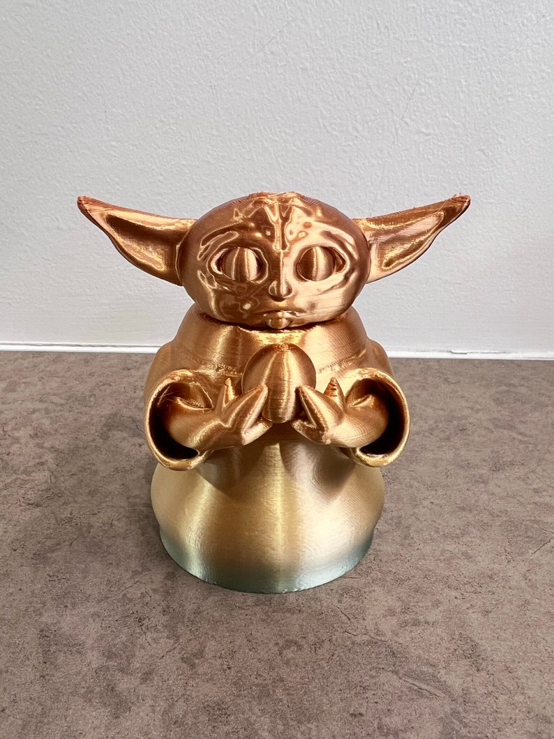 K&ouml;nnte beinhalten: Eine goldfarbene 3D-gedruckte Figur eines Charakters aus dem Star Wars-Franchise, bekannt als "The Child" oder "Baby Yoda". Die Figur ist mit ineinander verschr&auml;nkten H&auml;nden in einer gebets&auml;hnlichen Geste dargestellt.
