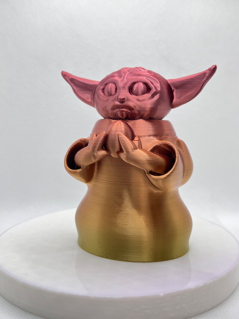 K&ouml;nnte beinhalten: Eine 3D-gedruckte Figur eines beliebten Charakters aus dem Star Wars-Franchise. Die Figur ist aus einem Farbverlaufsmaterial gefertigt, das von einer Ros&eacute;goldfarbe zu einer Goldfarbe &uuml;bergeht. Die Figur h&auml;lt ein kleines Objekt in ihren H&auml;nden.