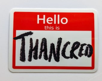 May include: A red and white name tag with the text "Hello, this is" and the name "Thancred" written in black marker.
