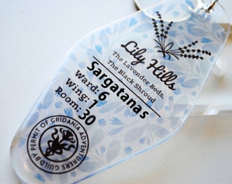 May include: Clear acrylic keychain with a blue floral pattern and black text. The text reads "Lily Hills, Sargatanas, Ward: 6, Wing: 1, Room: 30." A black emblem and the words "Permit of Gridania Adventurers Guild" are also visible.