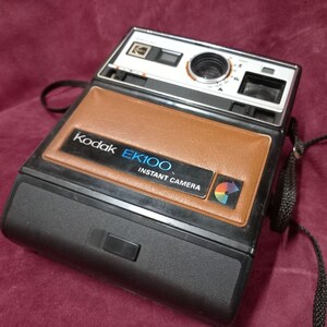 Puede incluir: Una cámara instantánea Kodak EK100 negra y marrón con una correa negra. La cámara tiene una cubierta de cuero marrón con las palabras "Kodak EK100 Instant Camera" impresas en ella.