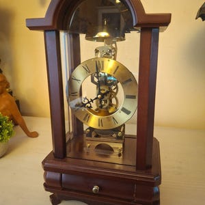 May include: A mantel clock with a dark brown wooden frame and a glass enclosure. The clock features a circular gold-toned face with black Roman numerals and intricate black hands. The internal brass gears are visible, and a small drawer is at the base.
