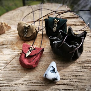 Könnte beinhalten: Eine Sammlung kleiner, handgefertigter Lederbeutel in verschiedenen Farben, darunter Braun, Rot und Grün, mit dekorativen Metallverschlüssen. Ein weißer und schwarzer Stein liegt daneben. Die Beutel liegen auf einer Holzoberfläche.