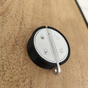 Puede incluir: Un mando a distancia negro y plateado con cuatro botones. Los botones tienen iconos para un candado, un desbloqueo, una campana y una casa.