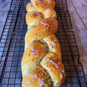 May include: A braided loaf of bread with a white glaze and colorful sprinkles on top. The bread is on a wire rack.