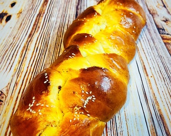 May include: A golden brown braided loaf of bread with sesame seeds on top, sitting on a wooden surface.