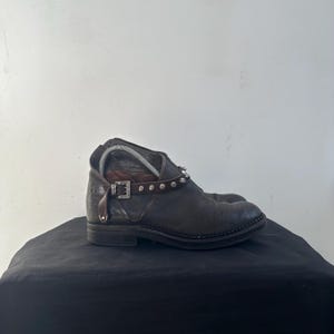 May include: Dark brown leather ankle boots with a low heel. The boots feature a studded strap around the ankle and a buckle detail. The boots are displayed on a black surface.