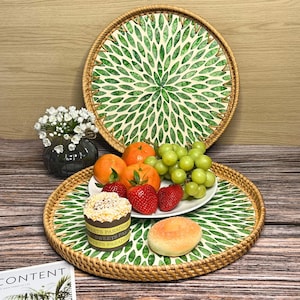 May include: Two round woven trays with green and white leaf designs. The smaller tray is filled with fruit, including oranges, grapes, and strawberries. A small cake sits on a white plate in the center of the tray.