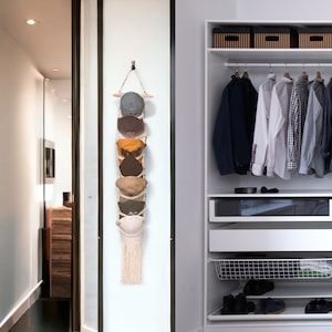 May include: A white macrame wall hanging with a wooden dowel and six baseball caps hanging from it. The caps are in various colors, including brown, yellow, and green.