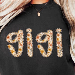 May include: Black t-shirt featuring the name "GIGI" in a floral design. The letters are filled with orange, yellow, and cream flowers, edged with a scalloped border. The shirt is a relaxed, comfortable style.