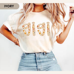 Op de afbeelding: Ivoor kleurig T-shirt met het woord "GIGI" in een bloemenontwerp. De letters zijn gevuld met oranje, gele en witte bloemen. Het shirt wordt gedragen met een blauwe jeans.