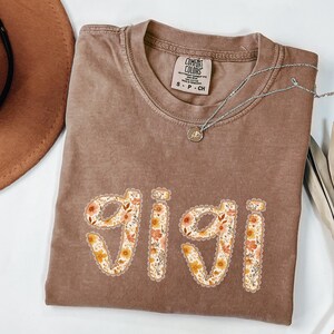 Op de afbeelding: Een bruin Comfort Colors t-shirt met de naam "Gigi" in een bloemenontwerp. De letters zijn gevuld met oranje, gele en witte bloemen. Een zilveren ketting met een kleine hanger is ook zichtbaar.