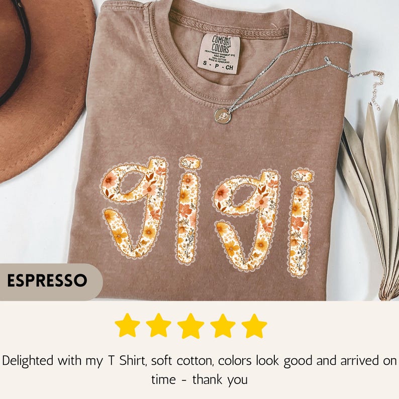 Op de afbeelding: Espresso-kleurig T-shirt met de naam "Gigi" in een bloemenontwerp. Het shirt heeft een ronde hals en korte mouwen. Een zilveren ketting met een kleine hanger rust op het shirt. Het shirt is gelabeld "Comfort Colors".