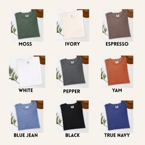 Op de afbeelding: Negen verschillend gekleurde t-shirts worden getoond, elk netjes opgevouwen. Kleuren zijn onder meer mos, ivoor, espresso, wit, peper, yam, spijkerbroekblauw, zwart en marineblauw. Elk shirt heeft een label en is op een neutrale achtergrond geplaatst.