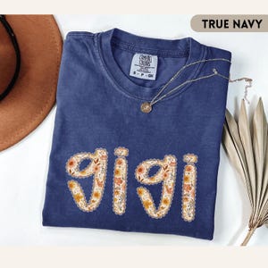 Op de afbeelding: Een marineblauw t-shirt met het woord "GIGI" in een bloemenontwerp. De shirt heeft een ronde hals en korte mouwen. Een gouden ketting met een hanger is over de shirt gedrapeerd. De shirt is op een witte ondergrond gevouwen.