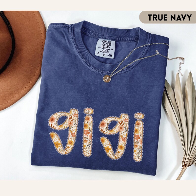Op de afbeelding: Een marineblauw t-shirt met het woord "GIGI" in een bloemenontwerp. De shirt heeft een ronde hals en korte mouwen. Een gouden ketting met een hanger is over de shirt gedrapeerd. De shirt is op een witte ondergrond gevouwen.