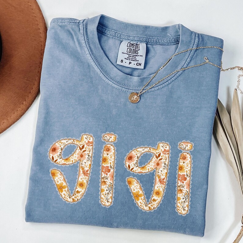Op de afbeelding: Een blauw Comfort Colors T-shirt met de naam "Gigi" in een bloemenontwerp. Het shirt heeft een ronde hals en korte mouwen. Een gouden ketting met een hanger is over het shirt gedrapeerd. Een bruine hoed is linksboven zichtbaar.