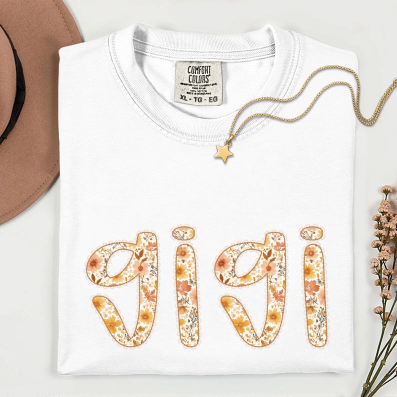 Op de afbeelding: Wit Comfort Colors T-shirt met de naam "Gigi" in een bloemenontwerp. Een gouden sterhanger ketting en een bruine hoed zijn ook zichtbaar. Het shirt is maat XL-TG-EG.