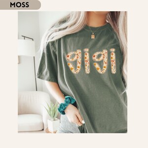 Op de afbeelding: Mosgroen t-shirt met het woord "Gigi" in een bloemenontwerp. De letters zijn gevuld met bloemen in geel, oranje en wit. Het shirt heeft korte mouwen en een relaxte pasvorm.