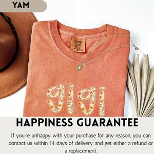 Op de afbeelding: Een koraalkleurig t-shirt met het woord "GIGI" geborduurd in een bloemenpatroon. Het shirt heeft een ronde hals en korte mouwen. Een gouden ketting met een hanger is over het shirt gedrapeerd. De tekst "HAPPINESS GUARANTEE" staat onderaan.