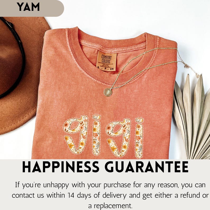 Op de afbeelding: Een koraalkleurig t-shirt met het woord "GIGI" geborduurd in een bloemenpatroon. Het shirt heeft een ronde hals en korte mouwen. Een gouden ketting met een hanger is over het shirt gedrapeerd. De tekst "HAPPINESS GUARANTEE" staat onderaan.