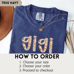 Op de afbeelding: Een marineblauw t-shirt met het woord "gigi" in een bloemenpatroon. De shirt heeft een ronde hals en korte mouwen. De tekst "TRUE NAVY" staat linksboven. De tekst "HOW TO ORDER" staat onderaan, met instructies.