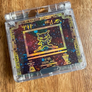 Può includere: Una console per videogiochi portatile Game Boy Color in plastica trasparente con un'immagine olografica di un gatto nero con occhi gialli. L'immagine è circondata da un bordo dorato e nero con il testo "HP HEAL" e "ITEM FIND" in un font stilizzato. La console ha il logo Nintendo nell'angolo in basso a destra.
