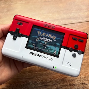 May include: A red, white, and black Game Boy Macro handheld console showing the Pokemon Emerald Version game. The console has a Nintendo logo and the text "GAME BOY MACRO" on the bottom.