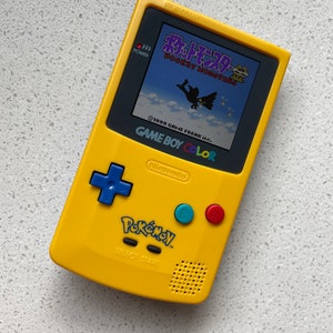 May include: A yellow Game Boy Color handheld game console with a screen displaying a Pokémon game. The console has a blue directional pad, red and blue buttons, and the Pokémon logo. The text "GAME BOY COLOR" and "Nintendo" are visible.