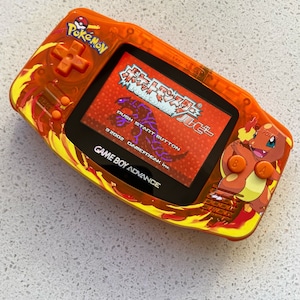 May include: An orange Game Boy Advance handheld console with a Pokémon theme. The console features a screen displaying a game interface with Japanese text. The design includes a Charmander graphic and flame-like patterns. The console has orange buttons and the Pokémon logo.