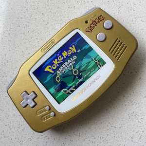 May include: A gold Game Boy Advance SP handheld gaming console with a white faceplate. The screen displays the Pokemon Emerald Version game. The console has a silver power button and a silver start and select button.