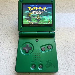 Puede incluir: Una consola de juegos portátil Game Boy Advance SP verde con una pantalla verde brillante que muestra la pantalla de título de la versión Pokemon Emerald. La consola tiene un D-pad negro, botones de cara y botones de selección e inicio.