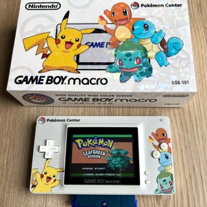 Puede incluir: Una consola Game Boy Macro blanca con una pantalla que muestra el juego Pokémon LeafGreen Version. La consola presenta a Pikachu y otros personajes Pokémon. La caja blanca tiene el logotipo de Nintendo y personajes Pokémon.