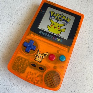 May include: An orange Game Boy Color handheld console displaying the Pokémon Yellow Pikachu game. The console has a blue directional pad, red and blue action buttons, and a Pikachu sticker. The screen shows the game's title and a yellow Pikachu character.