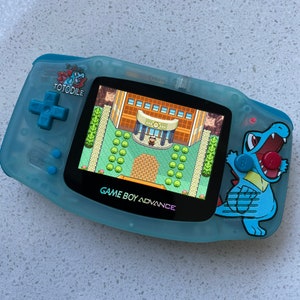 May include: A blue Game Boy Advance handheld gaming console with a clear shell and a  "TOTODILE" sticker on the front. The screen shows a pixelated image of a building with a character standing in front of it. The console has a blue and red button on the right side.